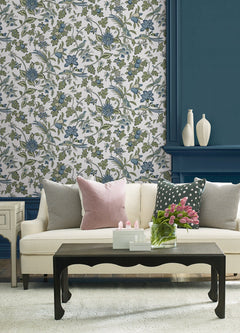 A cream sofa with pink and gray pillows sits in front of York Wallcoverings’ Parterre Jacobean Wallpaper. A black coffee table holds pink tulips in a glass vase, while white decorative vases are displayed on a blue shelf behind the sofa.
