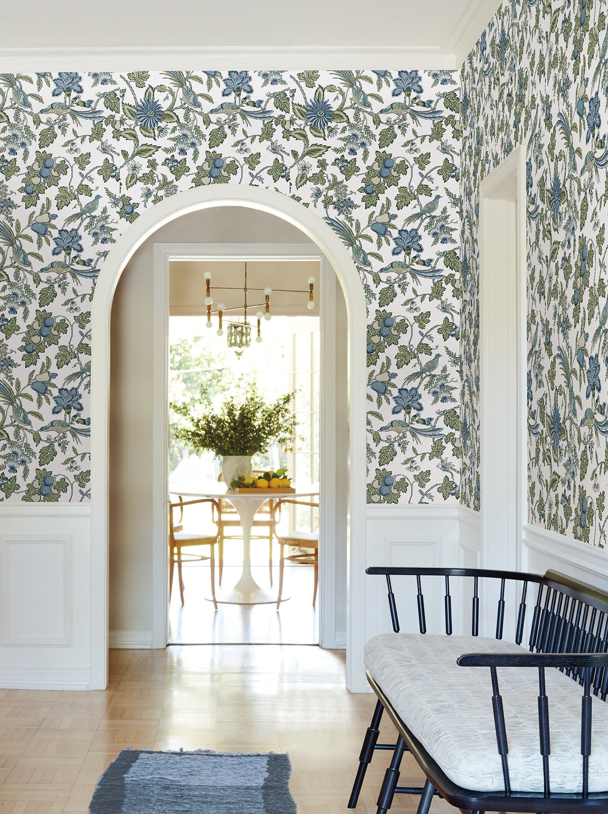 A room featuring Parterre Jacobean Wallpaper by York Wallcoverings in blue and green floral, paired with white trim, an arched doorway, a black bench with a cushion, and a view into a dining area furnished with a round table, chairs, and a vase of flowers.