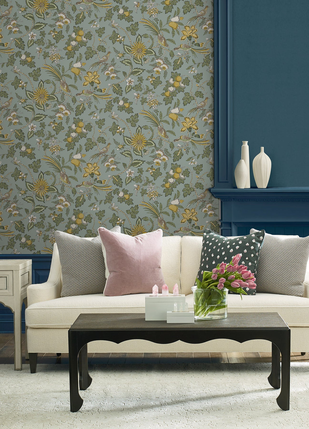 A cream sofa with pink and gray pillows sits in front of York Wallcoverings’ Parterre Jacobean Wallpaper and a blue wall. A black coffee table holds pink tulips and decor, while white vases are arranged on the mantel.