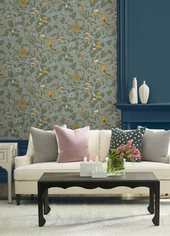 A cream sofa with pink and gray pillows sits in front of York Wallcoverings’ Parterre Jacobean Wallpaper and a blue wall. A black coffee table holds pink tulips and decor, while white vases are arranged on the mantel.