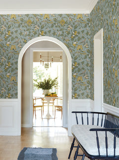 A hallway featuring Parterre Jacobean Wallpaper by York Wallcoverings in blue and yellow floral, complemented by white trim, a black bench with a cushion, and an arched doorway opening into a bright dining area with a round table and chairs.