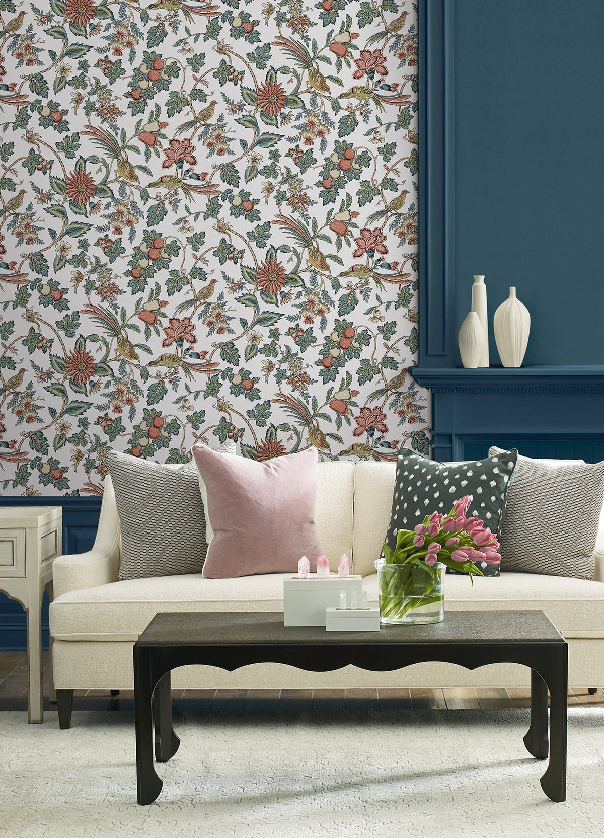 A living room featuring a cream sofa, pink and gray pillows, a black coffee table adorned with pink tulips, York Wallcoverings’ Parterre Jacobean Wallpaper, a blue paneled wall, and white decorative vases on the mantel.