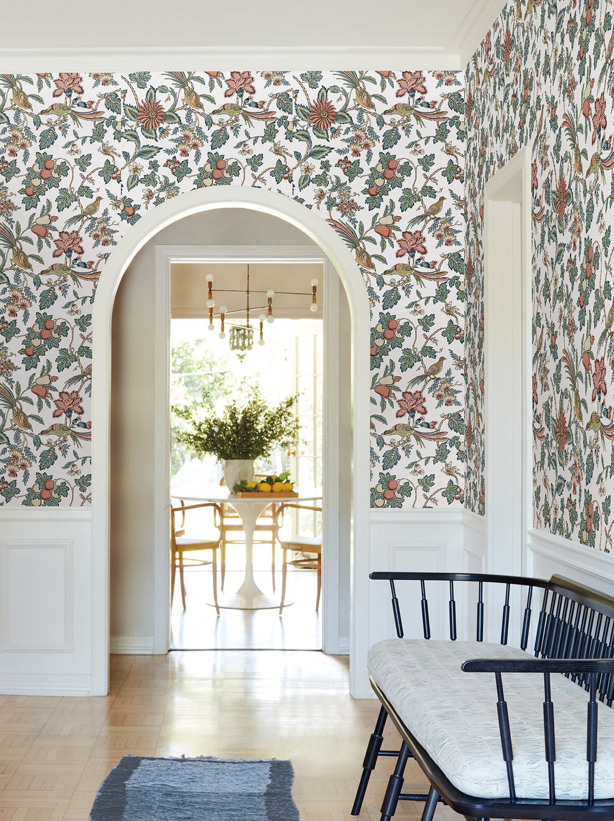 A hallway adorned with Parterre Jacobean Wallpaper by York Wallcoverings, featuring a black bench with a light cushion and a blue rug, leads through an arched doorway to a bright dining area with a round table, chairs, and a vase of greenery.