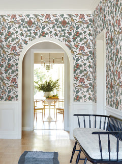 A hallway adorned with Parterre Jacobean Wallpaper by York Wallcoverings, featuring an arched doorway that leads to a dining area with a round white table, wooden chairs, a chandelier, and a vase of flowers; a black bench sits in the foreground.