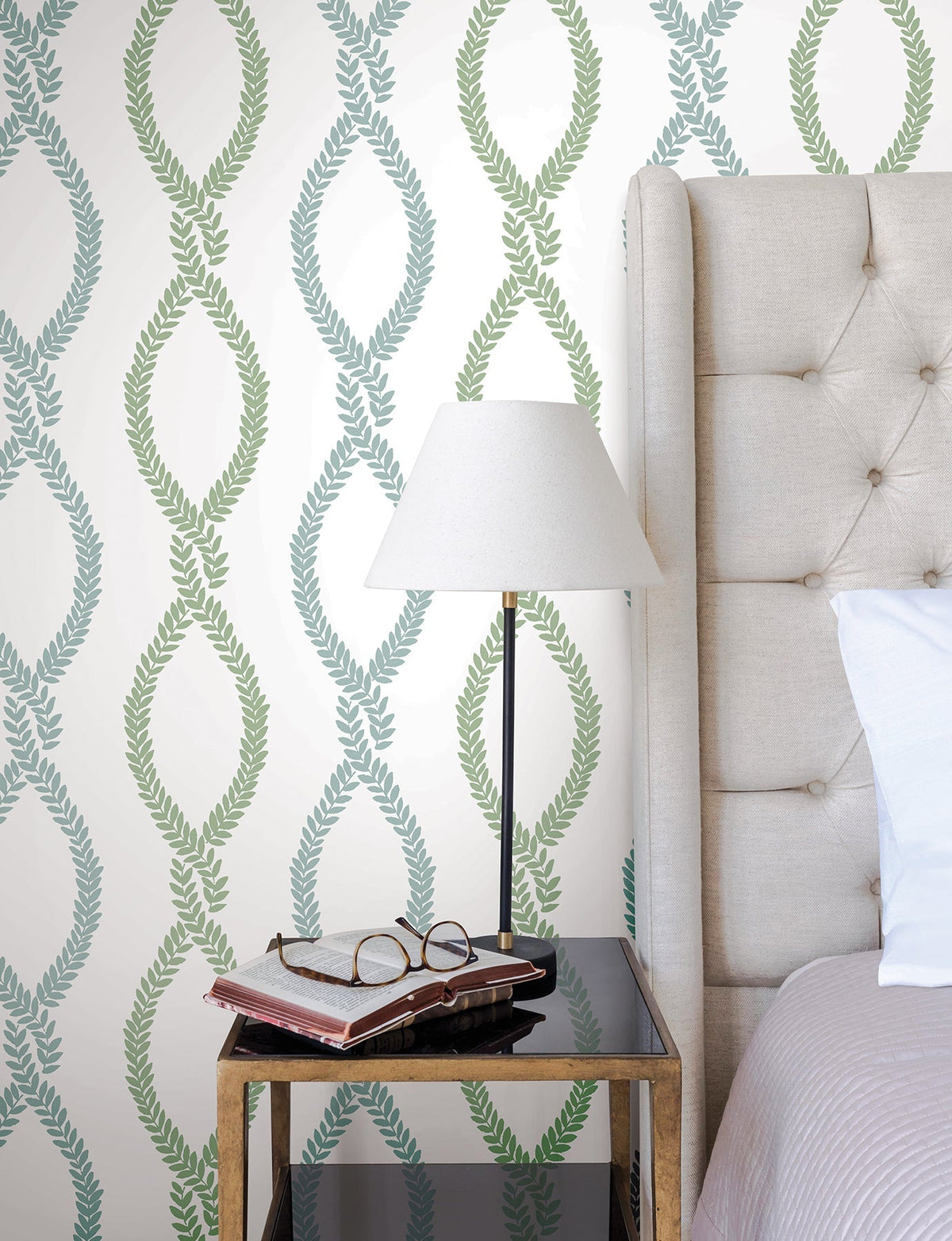 A black bedside table with books and eyeglasses sits next to a beige tufted bed. A lamp with a white shade rests on the table. The wall is adorned with York Wallcoverings’ Laureate Vine Trellis Wallpaper in a leafy green and blue pattern.