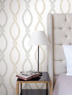 A beige upholstered bed, nightstand with books, eyeglasses, and a lamp. The walls are adorned with Laureate Vine Trellis Wallpaper by York Wallcoverings, featuring a repeating leaf pattern in soft tan and gray.