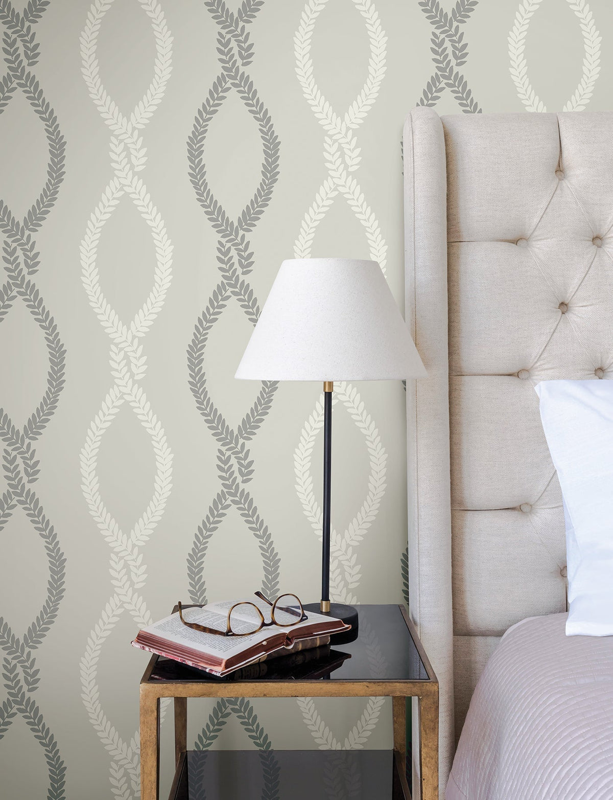 A bedside table with a lamp, books, and eyeglasses sits next to a tufted headboard. The wall features York Wallcoverings’ Laureate Vine Trellis Wallpaper, showcasing leafy oval designs in gray and cream.