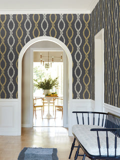 A hallway featuring Laureate Vine Trellis Wallpaper in a gray and gold pattern by York Wallcoverings, complemented by a black bench with a cushion, white wainscoting, and an archway that opens into a dining area furnished with a round table, chairs, and a vase of flowers.