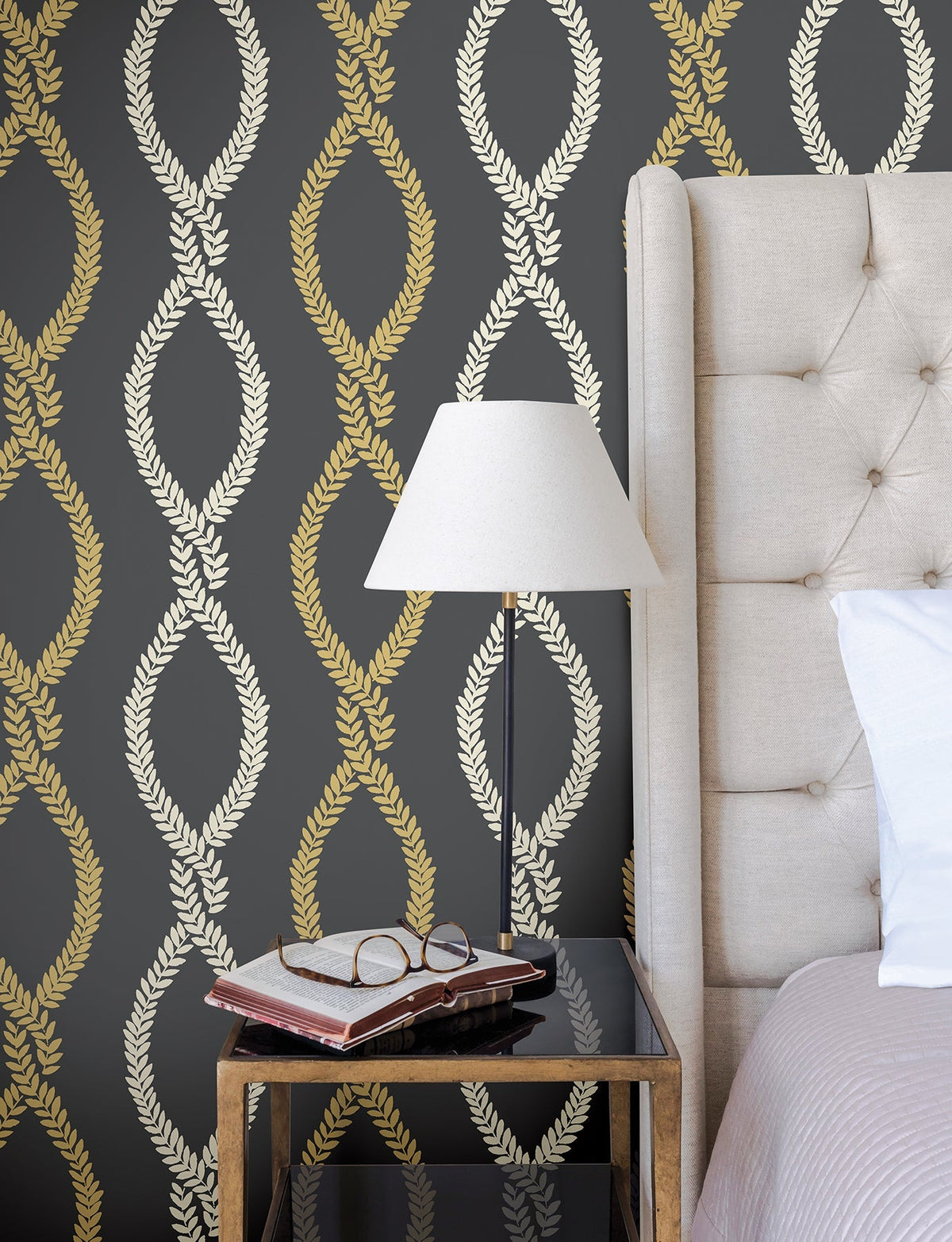 A beige bed with a tufted headboard sits beside a glass-top nightstand displaying books, glasses, and a lamp. The wall features York Wallcoverings’ Laureate Vine Trellis Wallpaper, showcasing an elegant gold and cream leaf pattern on a dark background.