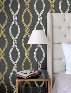 A beige tufted bed, black side table with books and glasses, and a white lamp sit against a dark wall covered in Laureate Vine Trellis Wallpaper by York Wallcoverings, featuring a gold and cream laurel wreath pattern.