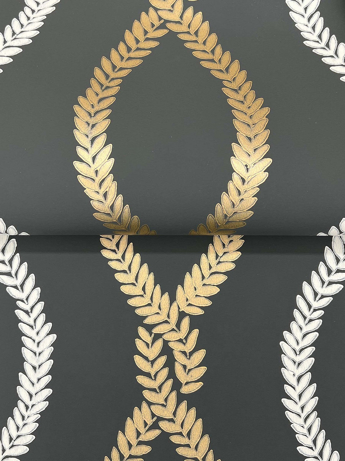 The Laureate Vine Trellis Wallpaper by York Wallcoverings features intersecting gold and silver laurel wreath motifs on a sophisticated dark gray background; the symmetrical design highlights a central gold wreath flanked by silver wreaths on each side.