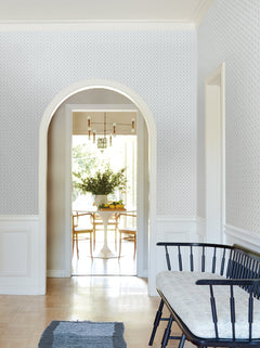 A bright hallway adorned with York Wallcoverings’ Priti Floral Grid Wallpaper features a black bench and leads to a dining room filled with sunlight, where a round white table, chairs, and a large vase of greenery await.