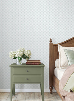 A green nightstand with two drawers, topped with a vase of white flowers and two stacked books, sits beside a wooden bed adorned with pillows, set against York Wallcoverings’ Priti Floral Grid Wallpaper and a light wood floor.