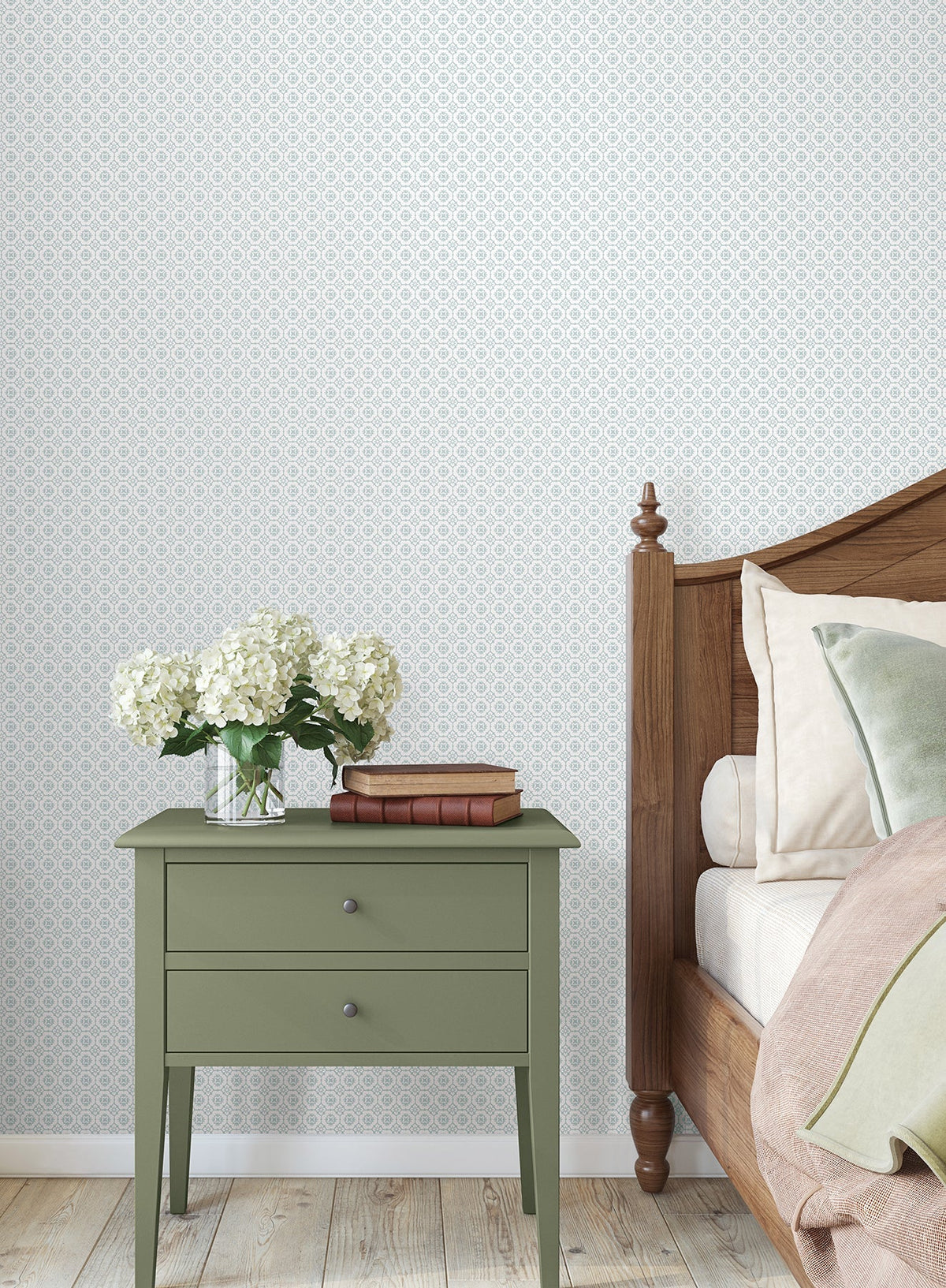 A green bedside table with two stacked books and a glass vase of white hydrangeas sits next to a wooden bed with beige and green bedding, set against York Wallcoverings’ Priti Floral Grid Wallpaper and a light wood floor.