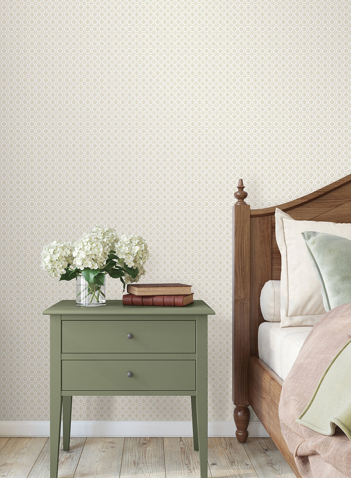 A green nightstand with two books and a vase of white flowers sits beside a wooden bed featuring cream and green bedding, set against Priti Floral Grid Wallpaper by York Wallcoverings, with a wooden floor underneath.