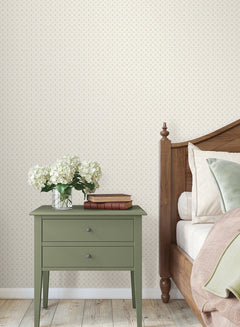 A green nightstand with two drawers sits next to a wooden bed adorned with pillows and a blanket. On the nightstand are two stacked books and a glass vase filled with white flowers. The room features Priti Floral Grid Wallpaper by York Wallcoverings and has a wooden floor.