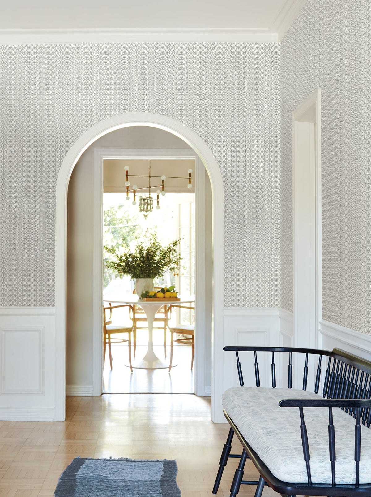 A bright hallway features Priti Floral Grid Wallpaper by York Wallcoverings, a black bench with a cushion, and a blue rug. Through a white archway, you can see a dining table with chairs and a flower arrangement, all illuminated by a chandelier.