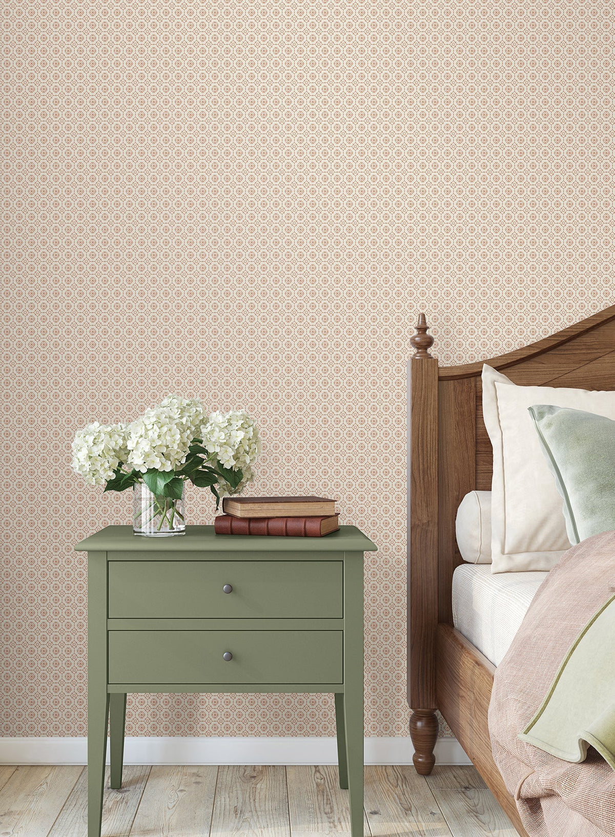A green nightstand with books and a vase of white flowers sits beside a wooden bed with beige bedding, set against York Wallcoverings’ Priti Floral Grid Wallpaper and a light wooden floor.