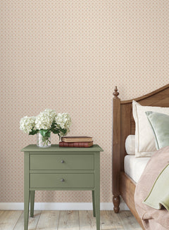 A green nightstand with books and a vase of white flowers sits beside a wooden bed with beige bedding, set against York Wallcoverings’ Priti Floral Grid Wallpaper and a light wooden floor.
