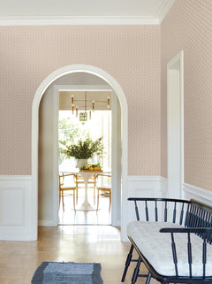 A hallway adorned with York Wallcoverings’ Priti Floral Grid Wallpaper and white trim leads through an arched doorway to a bright dining room with a round table, flowers, and chairs. A black bench with a cushion sits in the foreground.
