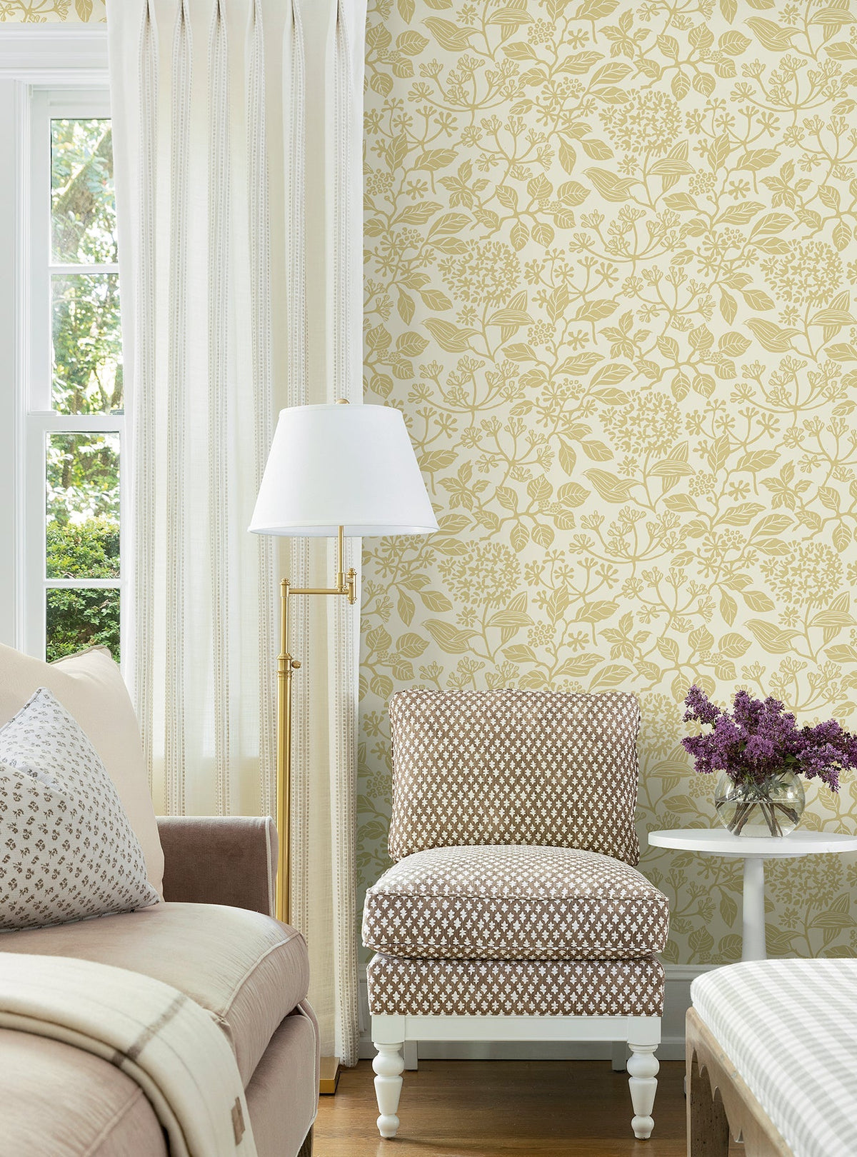 A cozy living room corner featuring Rowen Botanical Wallpaper by York Wallcoverings, a brown and white dotted chair, white floor lamp, light sofa, ottoman, and a table holding a vase of purple flowers near a window with cream curtains.
