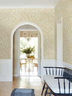 A hallway adorned with Rowen Botanical Wallpaper by York Wallcoverings, featuring a black bench with a gray cushion, offers a view through an archway into a dining room furnished with a white table, wooden chairs, and a vase of greenery. Natural light fills the rooms.