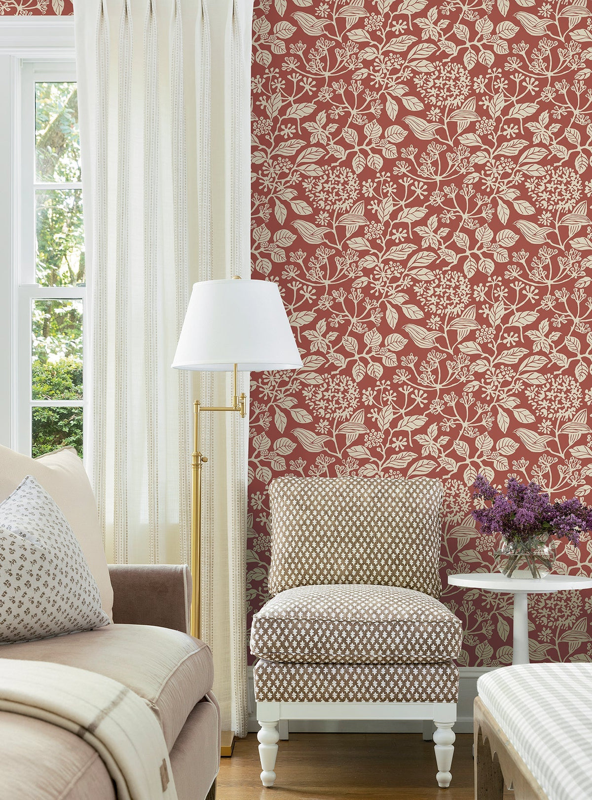 A living room with a beige sofa, patterned chair, white floor lamp, sheer curtains, and a small table holding a vase of purple flowers features the Rowen Botanical Wallpaper by York Wallcoverings in a red floral pattern.
