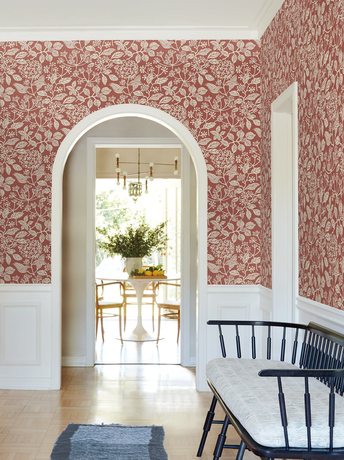 A hallway adorned with York Wallcoverings’ Rowen Botanical Wallpaper, accented by white trim, features a black bench with a cushion and an arched doorway that opens into a bright dining area furnished with a white table, wooden chairs, and a vase of greenery.