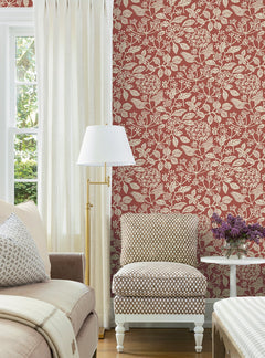 A living room features York Wallcoverings’ Rowen Botanical Wallpaper in a striking red and white floral design, complemented by a patterned armchair, beige sofa, white floor lamp, and white curtains. A small table with a vase of purple flowers sits nearby as sunlight streams in from the window.