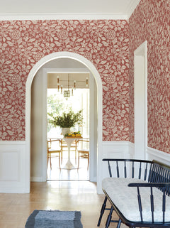 A hallway featuring Rowen Botanical Wallpaper by York Wallcoverings in red, complemented by white trim, an arched doorway, a black bench with a cushion, and a view into a bright dining room with a round table, chairs, and a vase of greenery.