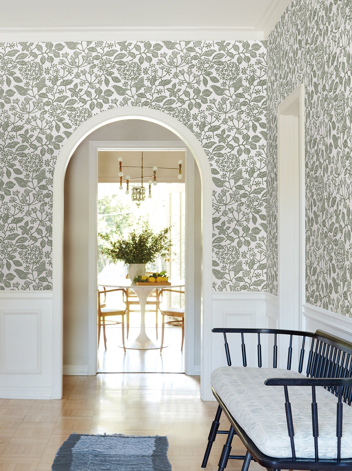 A hallway adorned with Rowen Botanical Wallpaper by York Wallcoverings features an arched doorway opening to a bright dining room with a round table and chairs, a bench topped with a cushion, and a small rug on the wooden floor.