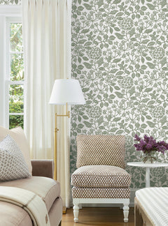 A cozy living room with a beige sofa, patterned armchair, gold floor lamp, Rowen Botanical Wallpaper by York Wallcoverings, cream curtains, and a small table with purple flowers by a window.
