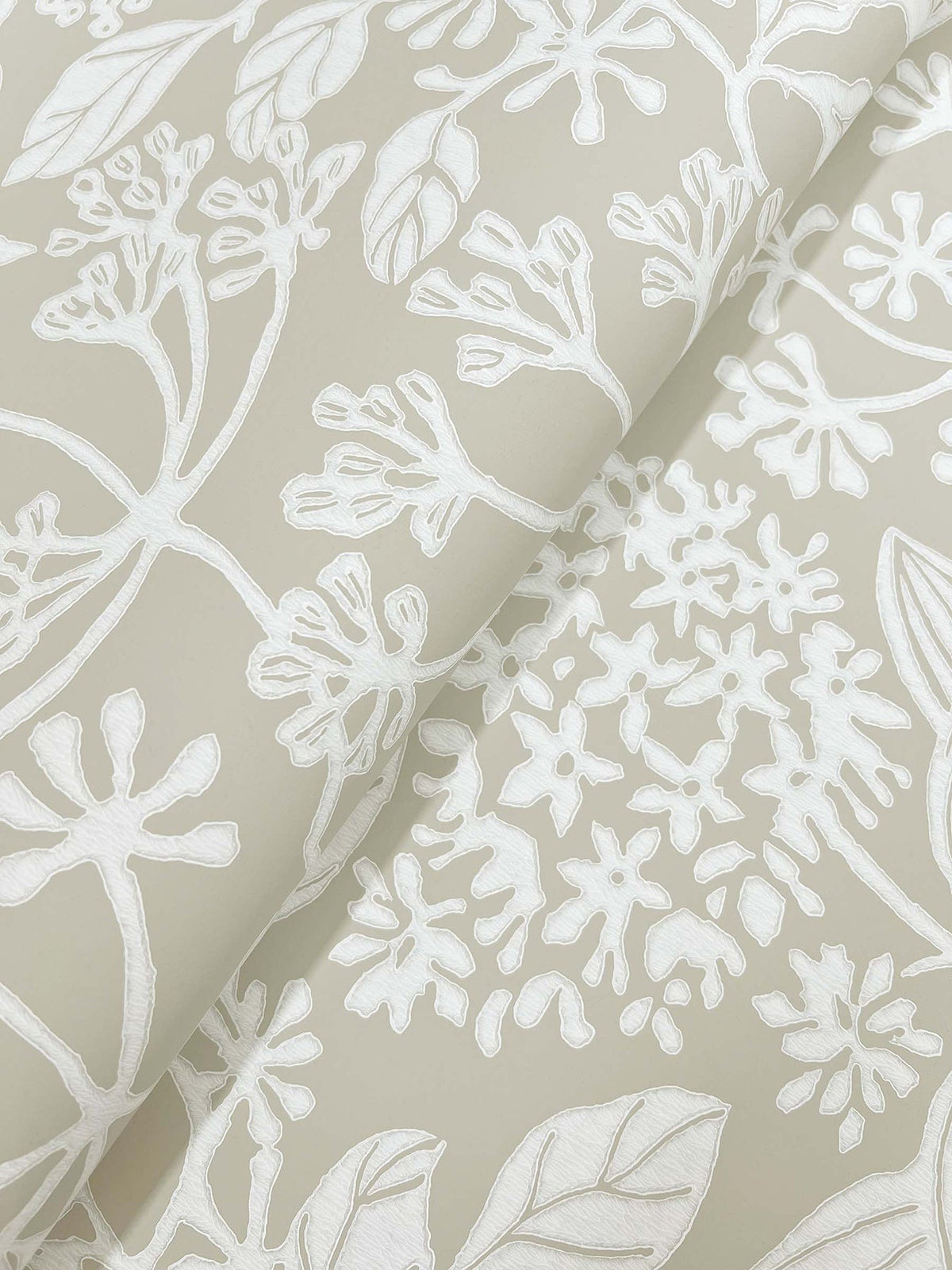 A close-up of Rowen Botanical Wallpaper by York Wallcoverings, featuring light beige tones with white, intricate floral and leaf patterns that create a textured, hand-drawn look. The wallpaper is shown slightly rolled to highlight its flexible nature.