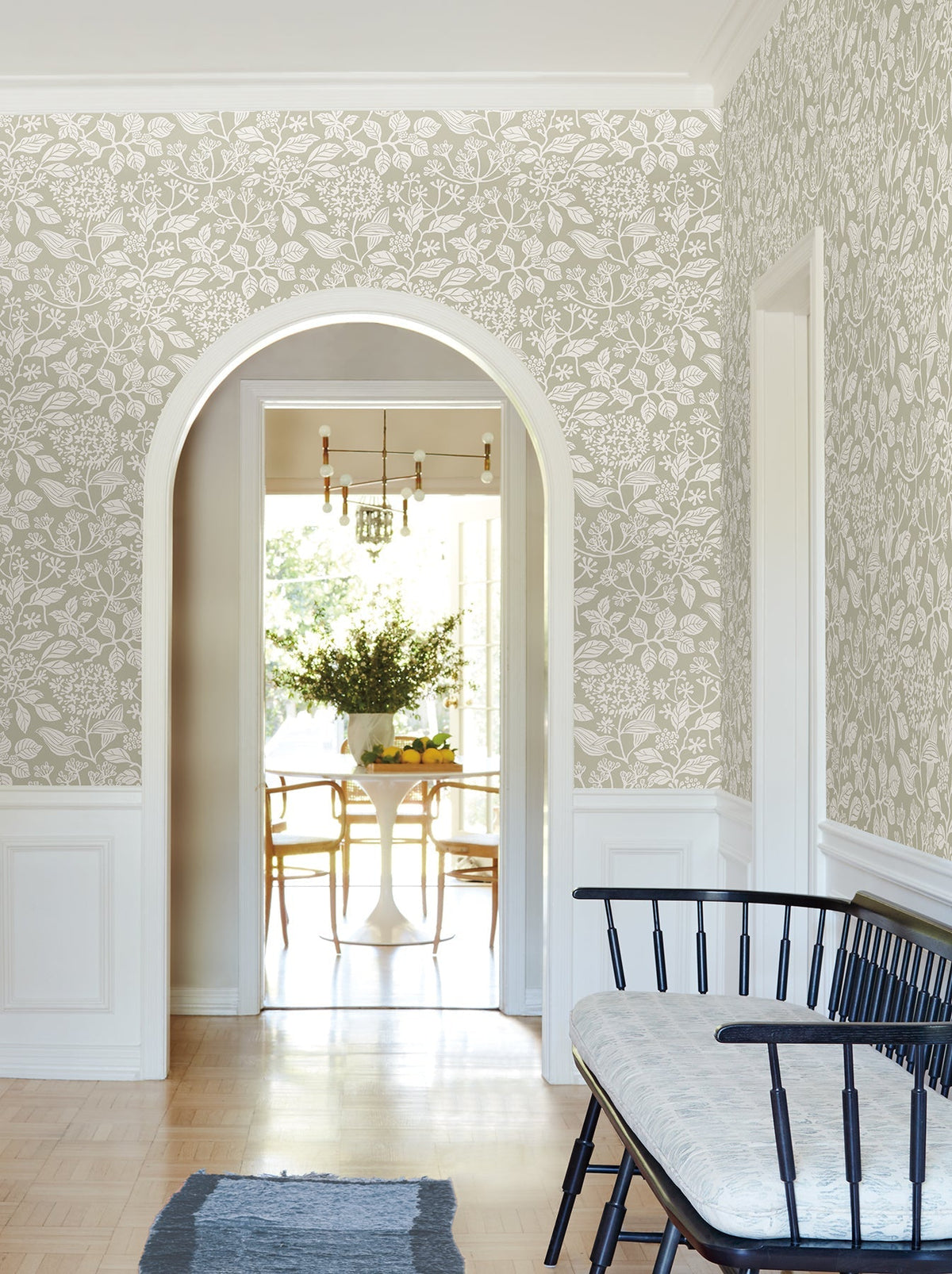 A hallway featuring Rowen Botanical Wallpaper by York Wallcoverings, with an arched doorway opening to a dining area furnished with a round table, wooden chairs, a black bench topped with a cushion, and a blue rug on light wood flooring.