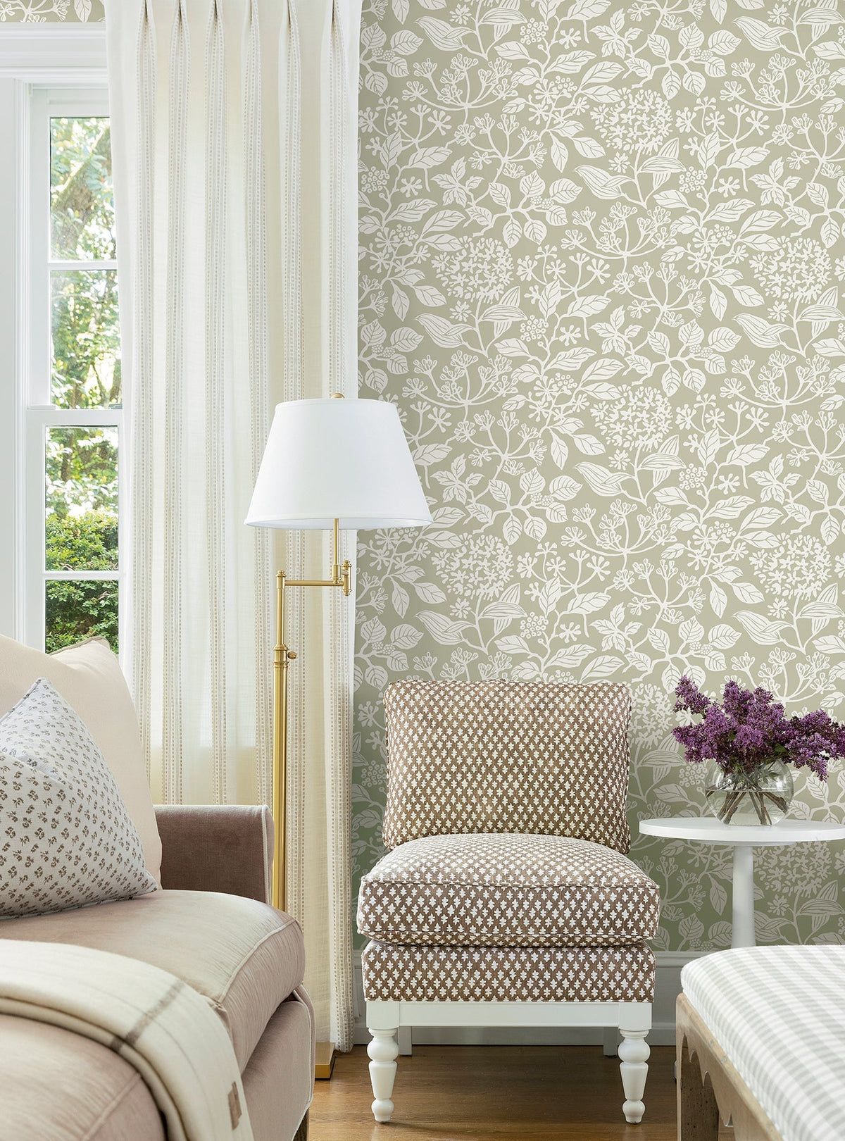 A cozy living room with Rowen Botanical Wallpaper by York Wallcoverings in beige, a patterned chair, light sofa, gold floor lamp, cream curtains, and a table holding a vase of purple flowers beside a sunny window.