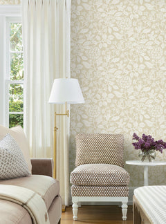 A cozy living room corner featuring a patterned armchair, York Wallcoverings’ Rowen Botanical Wallpaper, a white floor lamp, and a small round table with a vase of purple flowers beside a window dressed in cream curtains.