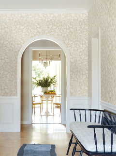 A hallway adorned with Rowen Botanical Wallpaper by York Wallcoverings features an arched doorway and a black bench with a cushion; in the background, the dining room showcases a white table, wooden chairs, fresh flowers, and streams of sunlight.
