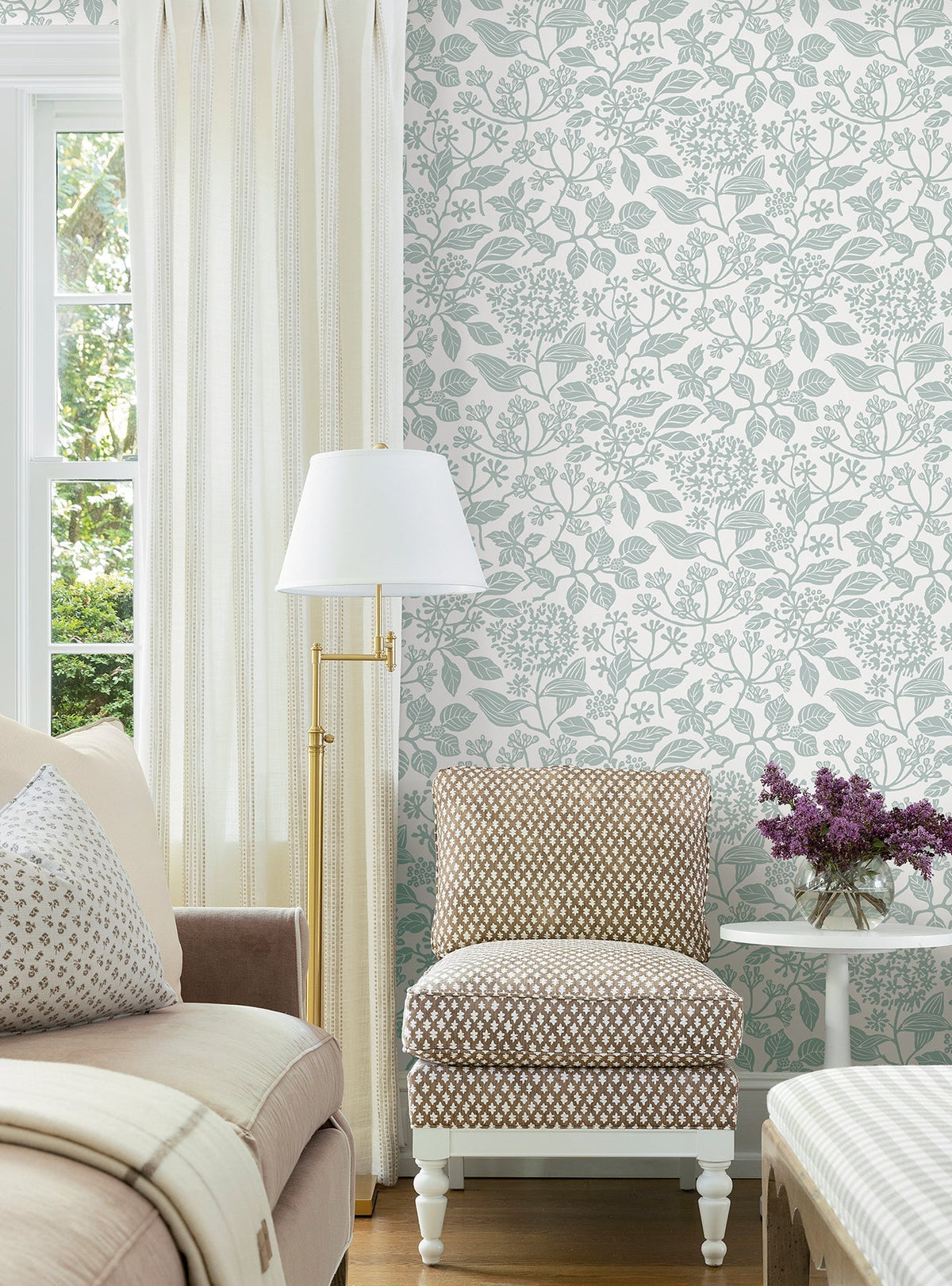 A cozy living room corner featuring a beige sofa, patterned accent chair, white side table with purple flowers, floor lamp, cream curtains, and York Wallcoverings’ Rowen Botanical Wallpaper.