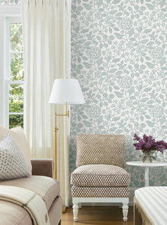 A cozy living room corner featuring a beige sofa, patterned accent chair, white side table with purple flowers, floor lamp, cream curtains, and York Wallcoverings’ Rowen Botanical Wallpaper.