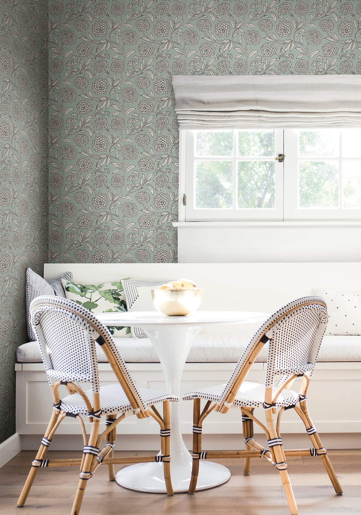 A cozy breakfast nook featuring a white round table, two woven chairs, a built-in bench with cushions, and a window with a Roman shade, set against York Wallcoverings’ Chatsworth Garden Floral Trail Wallpaper.