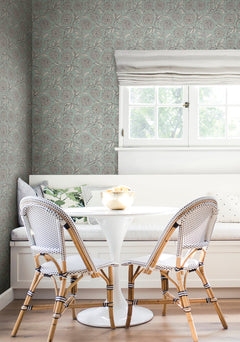 A cozy breakfast nook featuring a white round table, two woven chairs, a built-in bench with cushions, and a window with a Roman shade, set against York Wallcoverings’ Chatsworth Garden Floral Trail Wallpaper.