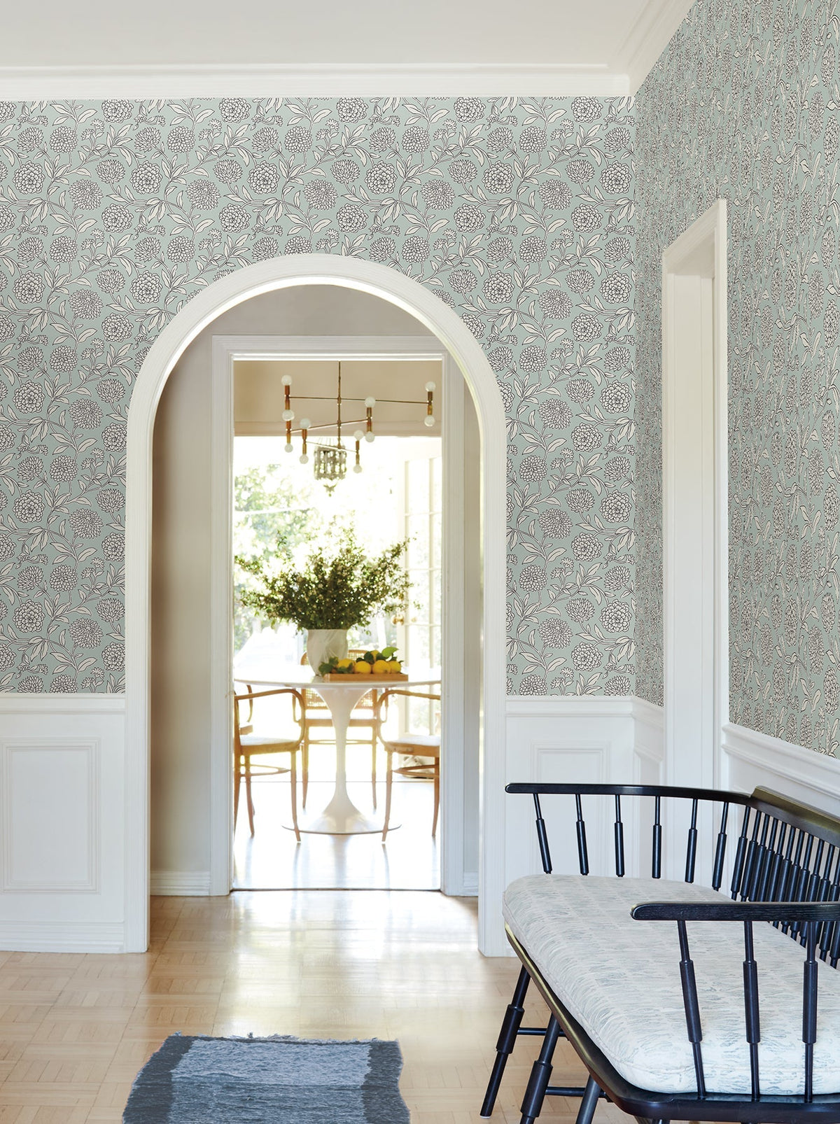 A view through an arched doorway shows a sunlit dining area with a round table, chairs, and a vase of flowers. The hallway features York Wallcoverings’ Chatsworth Garden Floral Trail Wallpaper, a cushioned bench, and a small rug on a light wood floor.
