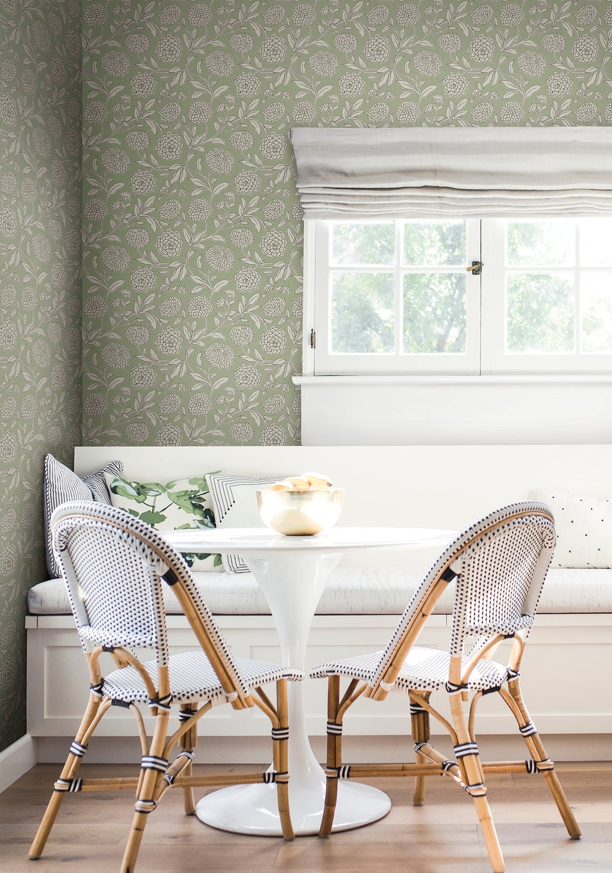 A small dining nook features a white round table, two woven chairs, and a built-in bench with pillows beneath a window. The walls are adorned with York Wallcoverings’ Chatsworth Garden Floral Trail Wallpaper in green. A bowl of fruit sits on the table.