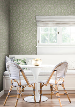 A cozy breakfast nook featuring a round white table, two woven chairs, a built-in bench with pillows, floral Chatsworth Garden Floral Trail Wallpaper by York Wallcoverings, and a window dressed with white roman shades letting in natural light.