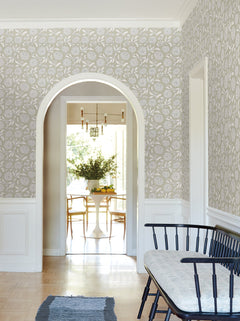 A hallway featuring York Wallcoverings’ Chatsworth Garden Floral Trail Wallpaper, a black bench with a cushion, an arched doorway, and a glimpse into a bright dining room with a round table, wooden chairs, and a vase of greenery.