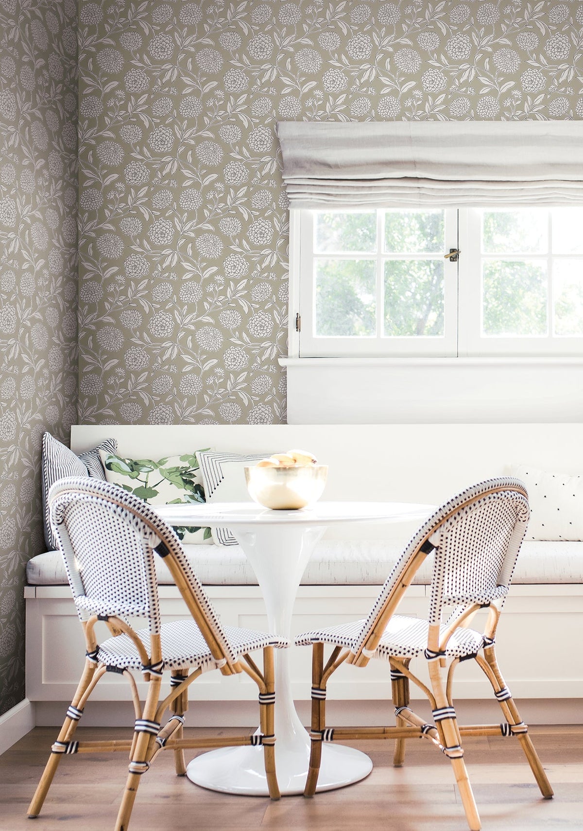 A bright breakfast nook features a white round table, two woven chairs, a built-in bench with cushions, and floral walls adorned with Chatsworth Garden Floral Trail Wallpaper by York Wallcoverings. Natural light streams in through a window with white blinds.