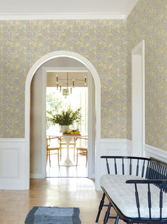 The bright hallway features Chatsworth Garden Floral Trail Wallpaper by York Wallcoverings in yellow and gray, complemented by crisp white trim, an arched doorway, and a black bench topped with a cushion. Through the archway, there’s a view into a dining room furnished with a round table, chairs, and a vase of greenery.