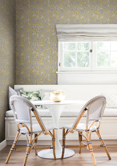 A bright nook featuring a white round table, two woven chairs, a built-in bench with pillows, and a window with a white shade that lets in natural light—all set against York Wallcoverings’ Chatsworth Garden Floral Trail Wallpaper.