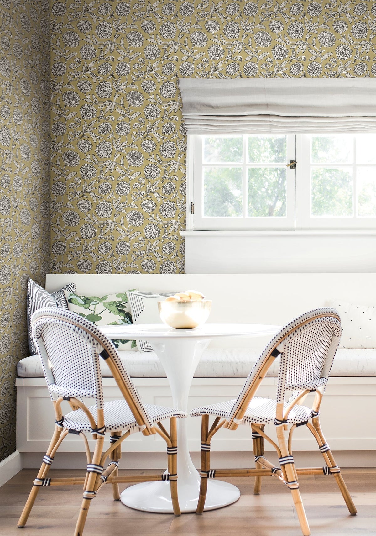 A cozy breakfast nook with a round white table, two wicker chairs, a built-in bench with cushions, and a window above. The walls are covered in Chatsworth Garden Floral Trail Wallpaper by York Wallcoverings.