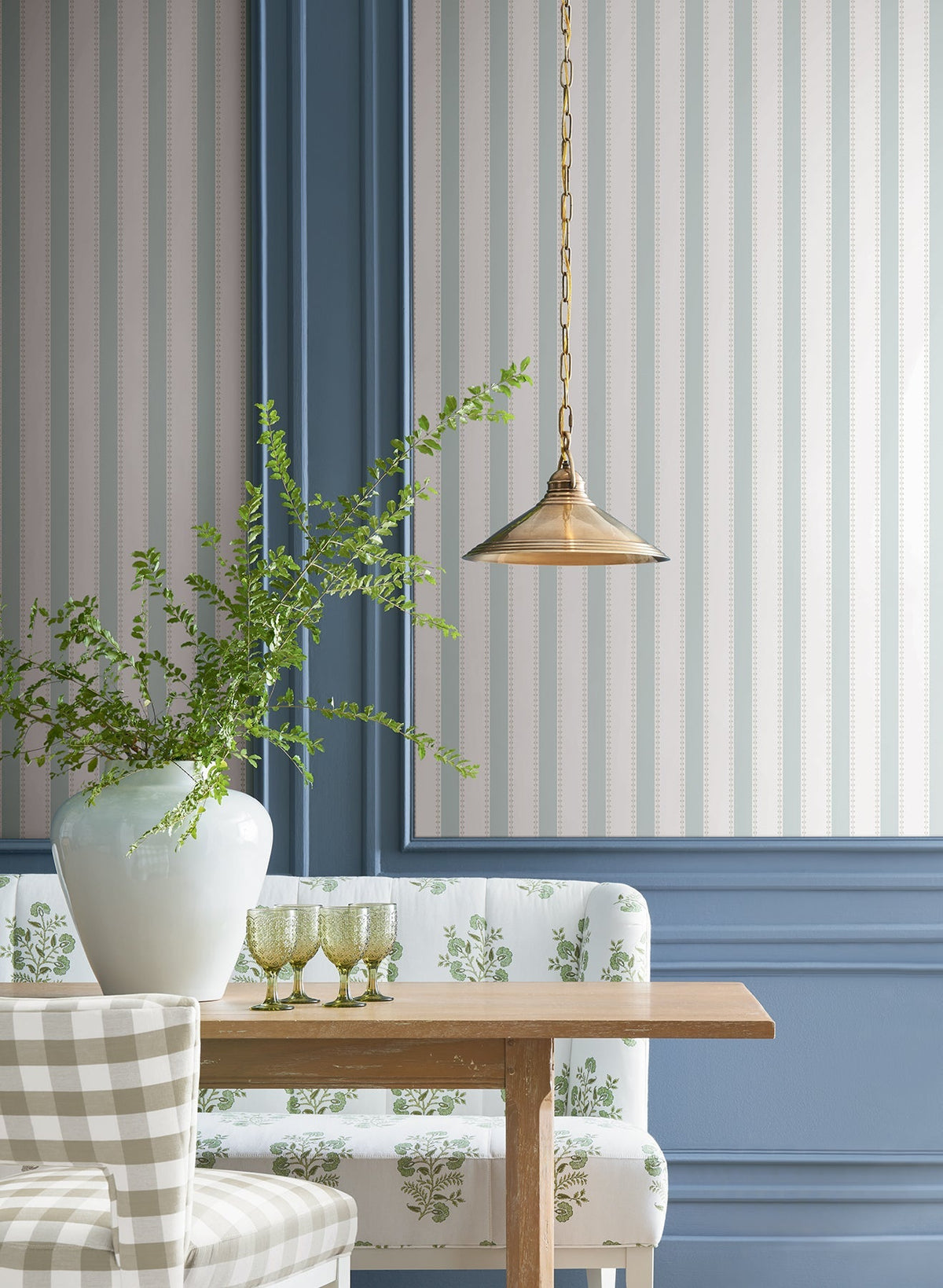 A dining nook features a wooden table, a patterned upholstered bench, a plaid chair, green glasses, and a large white vase with greenery. The walls are decorated with blue paneling and Tilford Awning Stripe Wallpaper by York Wallcoverings, while a brass pendant light hangs above.